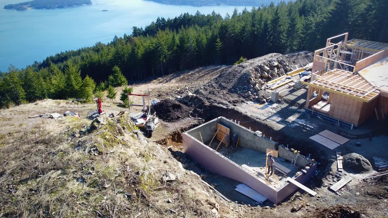 Aerial view of a construction site on a hillside overlooking a body of water, featuring the early stages of a building structure with forest surroundings.